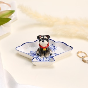 May include: A fan-shaped ceramic trinket dish featuring a blue floral pattern on a white speckled base. A small, three-dimensional black and white dog figurine with a red collar and a yellow bell is attached to the centre of the dish.