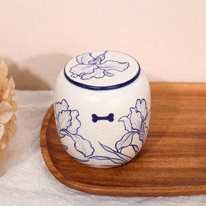 May include: A white ceramic jar with a lid, featuring blue hand-painted iris flower illustrations. The front of the jar displays a small blue bone icon above the embossed name Jasper. The jar has a rounded shape and a smooth, glossy finish.