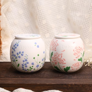 May include: Two round white ceramic jars with lids. The left jar features blue floral patterns and the name Archie embossed on the front. The right jar displays pink chrysanthemum-style flowers and the name Toby embossed on the front surface.