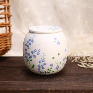 May include: A round white ceramic urn with a matching lid, featuring hand-painted blue forget-me-not flowers with green stems and leaves. The name Archie is embossed in the center of the vessel, which has a smooth matte finish and a spherical shape.