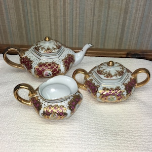 May include: A white tea set with gold trim and pink floral designs. The set includes a teapot, creamer, and sugar bowl.
