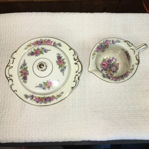 May include: A vintage porcelain butter dish with a lid and a matching creamer. The dish and creamer are decorated with a floral pattern in shades of pink, purple, and green. The edges of the dish and creamer are trimmed with gold.