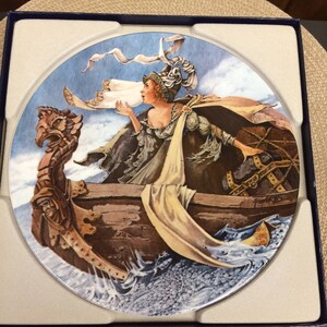 May include: A round ceramic plate with a colorful illustration of a woman in a boat with a sail. The woman is blowing on the sail to make it move. The boat is on a blue and white sea with waves. The plate is in a white box with a blue border.