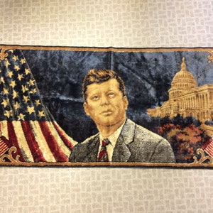 May include: A tapestry featuring a portrait of John F. Kennedy, the American flag, and the US Capitol building. The tapestry is made of a dark blue fabric with a velvet-like texture.