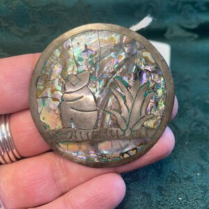 May include: Round, silver-toned pendant with an abalone shell inlay. The inlay features a carved design of a creature sitting on a branch with foliage. The pendant is approximately 2 inches in diameter.
