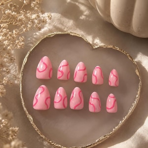 May include: A set of ten press-on nails in a light pink base color with a darker pink wavy line pattern. The nails are arranged in two rows of five on a heart-shaped tray with a gold-colored rim, featuring an almond-shaped tip design.