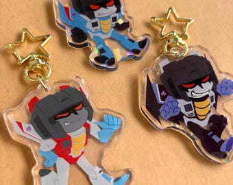 Op de afbeelding: Drie doorzichtige acryl sleutelhangers met cartoon robotfiguren met zwarte helmen en rode ogen. Elke sleutelhanger heeft een goudkleurige stervormige sluiting. De figuren zijn afgebeeld in dynamische houdingen met blauwe, rode en zwarte kleuren.