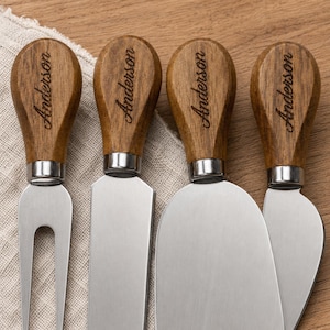 May include: A set of four cheese knives with stainless steel blades and wooden handles, each engraved with the name Anderson. The set includes a two-pronged fork, a rectangular spreader, a wide spade, and a pointed knife, all featuring silver ferrules.