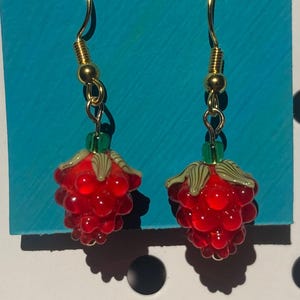 May include: A pair of dangle earrings featuring red glass raspberry charms with green leaves, suspended from gold-toned metal hooks. The earrings are displayed on a small, rectangular teal card with a single hole at the top center.