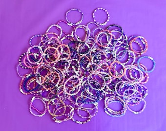 Puede incluir: Una gran colección de pulseras circulares de cuentas dispuestas en un grupo. Cada pulsera está compuesta por pequeñas cuentas multicolores en tonos rosa, morado, azul y blanco, algunas con pequeñas cuentas de letras blancas.
