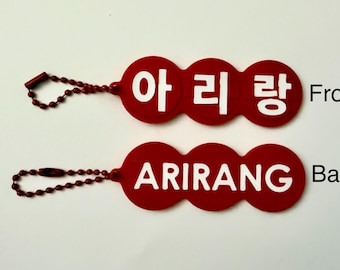 May include: Two red keychain tags with a triple-circle shape and a red ball chain. The top tag features the white Korean text 아리랑, while the bottom tag displays the white English text ARIRANG. The words Front and Back are printed in black.