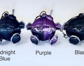 Puede incluir: Tres llaveros esféricos con forma de personaje, extremidades pequeñas y una antena superior. El llavero de la izquierda está etiquetado como Midnight Blue, el central como Purple y el de la derecha como Black. Cada uno tiene una costura central.