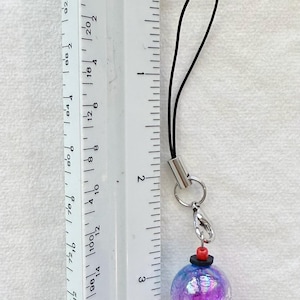 Puede incluir: Un colgante para tel&eacute;fono con una cuenta esf&eacute;rica iridiscente de color morado y azul con textura brillante, unido a un cord&oacute;n negro. Una peque&ntilde;a cuenta roja y un disco negro se sit&uacute;an sobre la esfera, con una base negra debajo. Mide 10,16 cm.