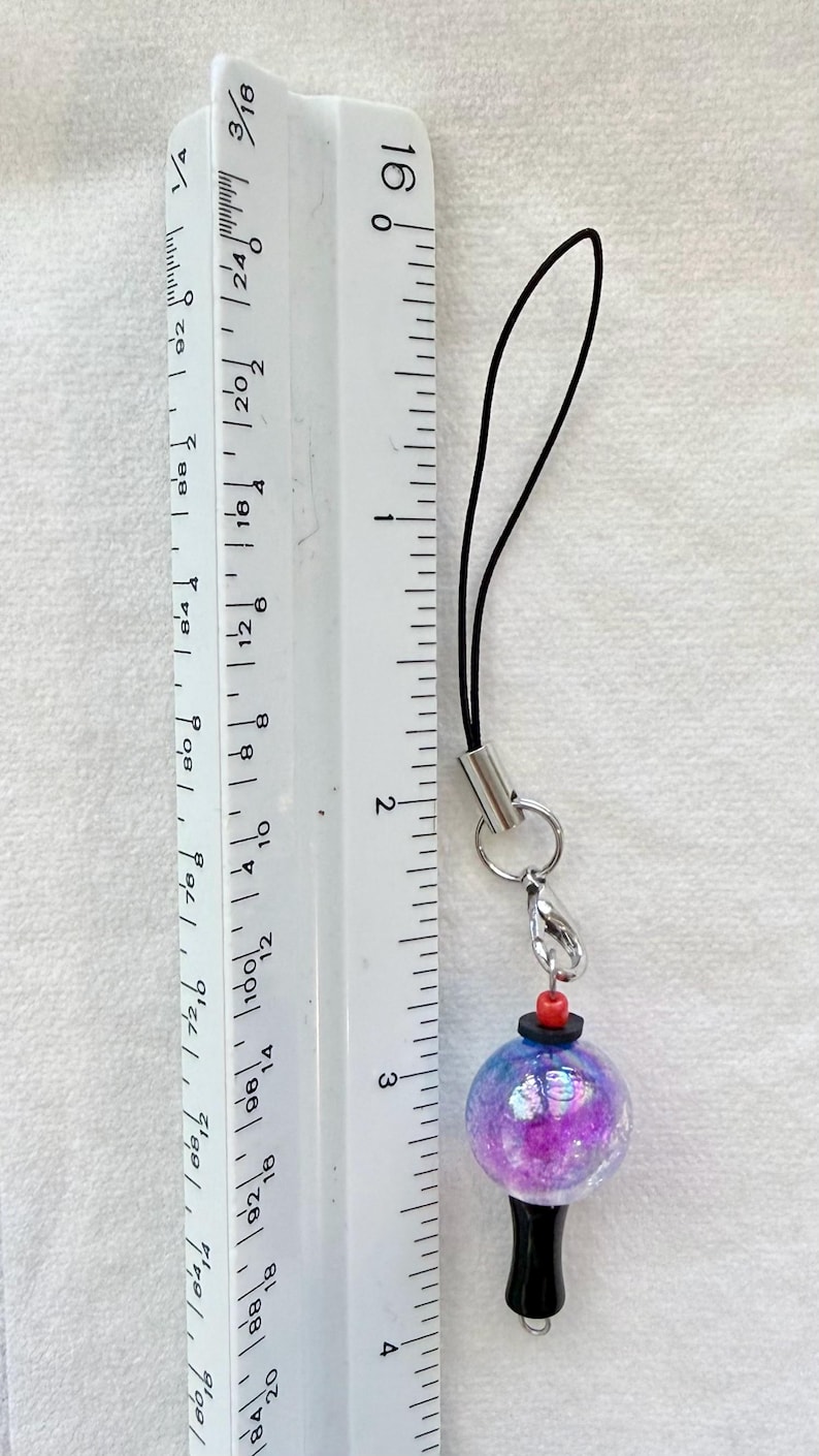 Puede incluir: Un colgante para tel&eacute;fono con una cuenta esf&eacute;rica iridiscente de color morado y azul con textura brillante, unido a un cord&oacute;n negro. Una peque&ntilde;a cuenta roja y un disco negro se sit&uacute;an sobre la esfera, con una base negra debajo. Mide 10,16 cm.