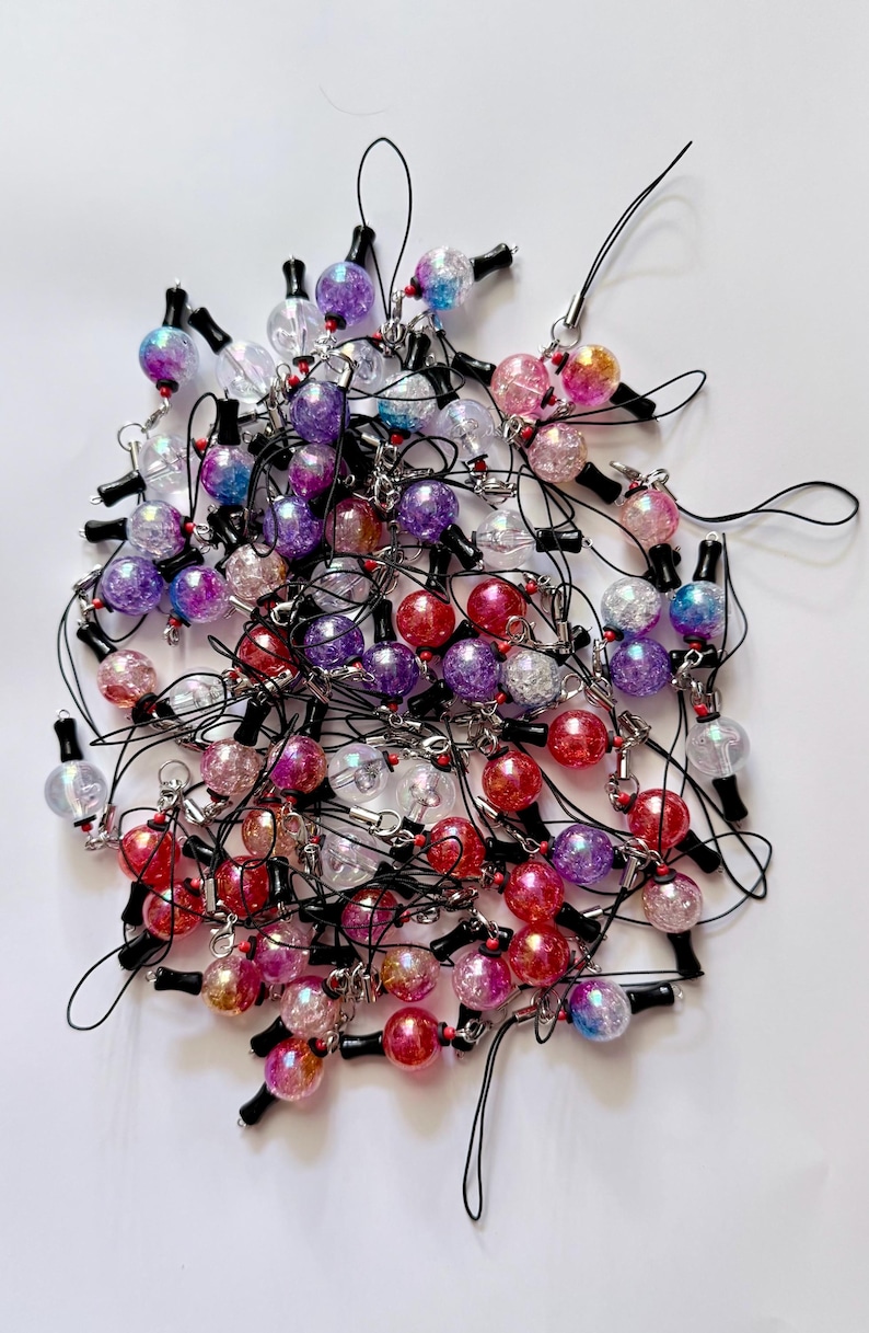 Puede incluir: Una colecci&oacute;n de numerosos dijes esf&eacute;ricos con textura agrietada y acabado iridiscente en tonos rojo, morado, rosa, azul y transparente. Cada dije est&aacute; unido a un lazo de cord&oacute;n negro con un peque&ntilde;o conector de metal de tono plateado.