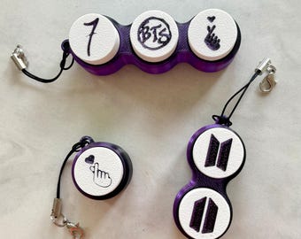May include: Three purple and white plastic keychains with black lanyards and metal clasps. One keychain has three circular white discs with the number 7, the BTS logo, and a finger heart. Another is a single disc with a finger heart, and the third has two discs with the BTS logo.
