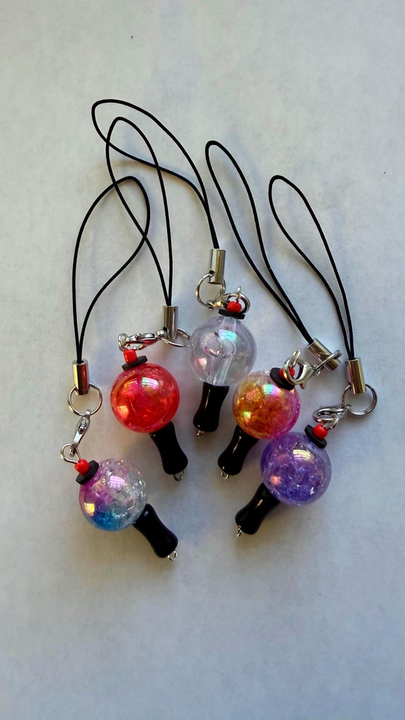 Puede incluir: Cinco colgantes para tel&eacute;fono con cuentas esf&eacute;ricas de acabados iridiscentes y brillantes en rojo, transparente, naranja, morado y azul-morado. Cada colgante tiene un mango negro, un peque&ntilde;o detalle de cuenta roja, una anilla de metal plateado y un cord&oacute;n negro.