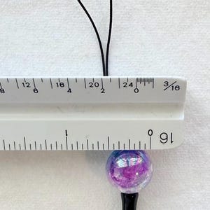 Puede incluir: Un dije esf&eacute;rico con acabado p&uacute;rpura e iridiscente unido a un cord&oacute;n negro. Una regla blanca se coloca sobre el dije, mostrando que la esfera tiene un di&aacute;metro de aproximadamente 1,9 cm. El dije cuenta con un componente de base negro.