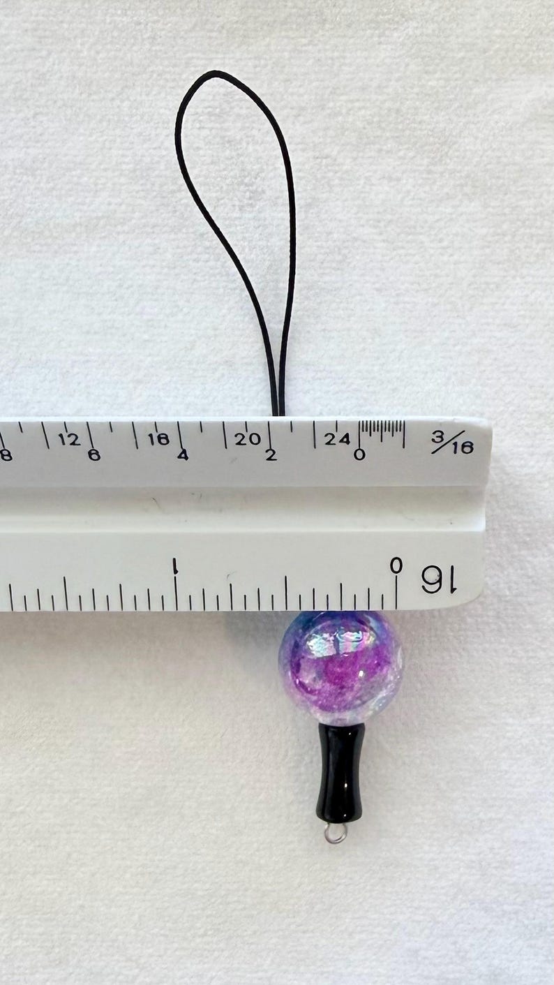 Puede incluir: Un dije esf&eacute;rico con acabado p&uacute;rpura e iridiscente unido a un cord&oacute;n negro. Una regla blanca se coloca sobre el dije, mostrando que la esfera tiene un di&aacute;metro de aproximadamente 1,9 cm. El dije cuenta con un componente de base negro.