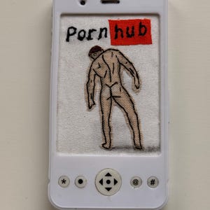 May include: A white plastic frame shaped like a smartphone containing a piece of white fabric with black hand-embroidered text reading "Porn hub" with a red rectangle, and a black-outlined embroidered figure of a person standing with their back turned.