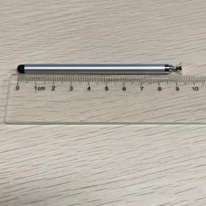 以下が含まれることがあります： 黒いゴム製の先端が付いた銀色のスタイラスペンが、透明なプラスチック製の定規の上に置かれています。定規の目盛りから、スタイラスペンの長さは約9センチメートルであることがわかります。スタイラスペンは金属光沢のある仕上げで、端には小さな円形のパーツが付いています。