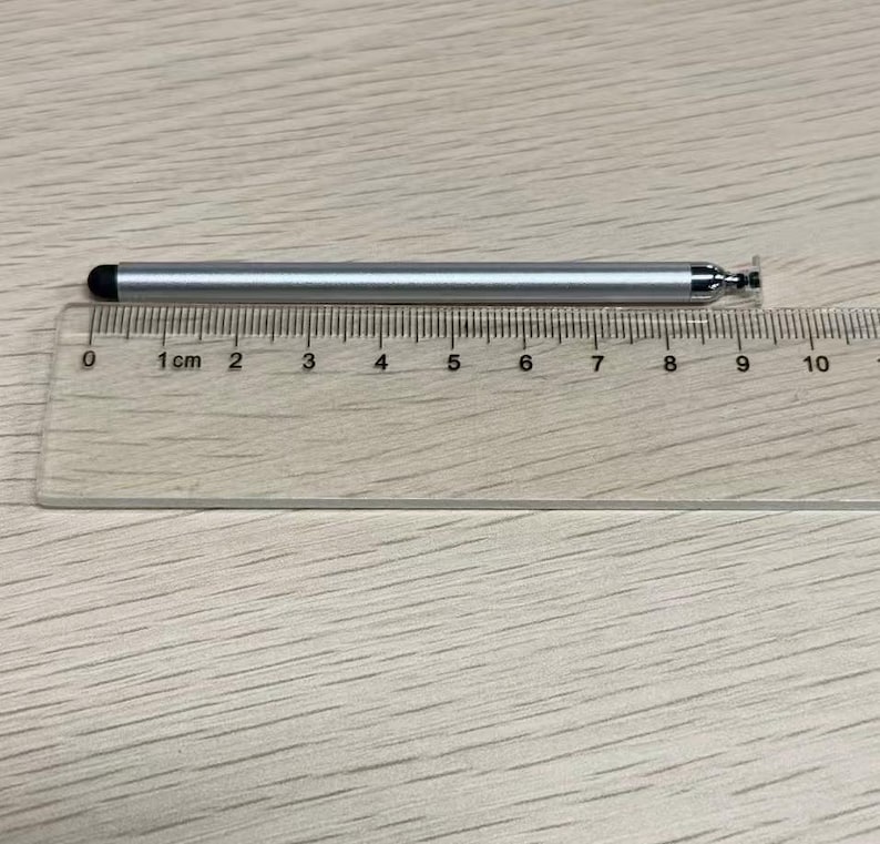 以下が含まれることがあります： 黒いゴム製の先端が付いた銀色のスタイラスペンが、透明なプラスチック製の定規の上に置かれています。定規の目盛りから、スタイラスペンの長さは約9センチメートルであることがわかります。スタイラスペンは金属光沢のある仕上げで、端には小さな円形のパーツが付いています。