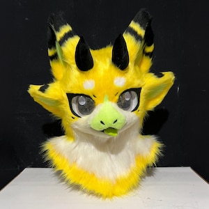 May include: A bright yellow and white faux fur animal mask featuring large black-tipped ears, two small black horns, and wide eyes with black outlines. The creature has a light green nose and a small green tongue protruding from its mouth.