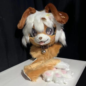 May include: A plush animal head mask featuring long brown and white fur, large eyes with heart-shaped pupils, and a small pink tongue. The mask includes a brown collar with a silver bell and matching furry hand paws with pink paw pads.