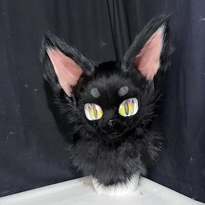 May include: A black faux fur animal head mask featuring large, pointed ears with pink inner lining, prominent yellow and orange eyes with vertical pupils, and small grey patches above the eyes. The mask has a dense, textured black fur covering.