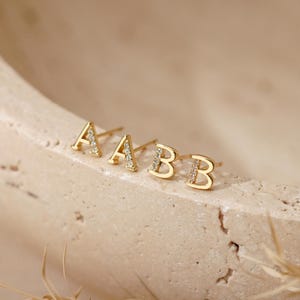 May include: Four gold stud earrings shaped like the letters A and B. Each letter features a vertical line of small, clear, sparkling stones embedded along one side of the character. The earrings are arranged in a row on a curved, textured surface.