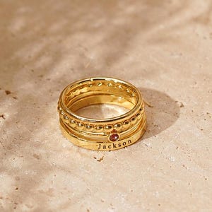 May include: A stack of three gold-toned rings. The top ring features a beaded texture, the middle ring has a small embedded red stone, and the bottom ring is a smooth band engraved with the name Jackson in a simple black sans-serif font.