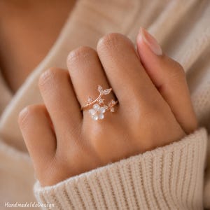 May include: A rose gold ring featuring a delicate branch design adorned with small clear stones and a single white flower-shaped accent. The band is open-ended, showcasing a floral motif that wraps around the finger with sparkling details.