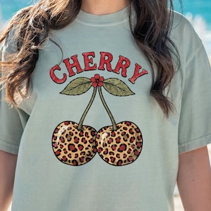 Op de afbeelding: Een licht saliegroen t-shirt met een grafische print van twee kersen met een luipaardpatroon. Boven de kersen staat het woord CHERRY in rode hoofdletters gedrukt met een klein rood bloemetje tussen de twee groene bladeren.
