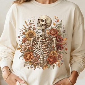 May include: A cream-colored long-sleeve sweatshirt featuring a central graphic of a human skeleton torso surrounded by a vibrant arrangement of autumn-toned flowers, including sunflowers, poppies, and ferns in shades of orange, red, and brown.