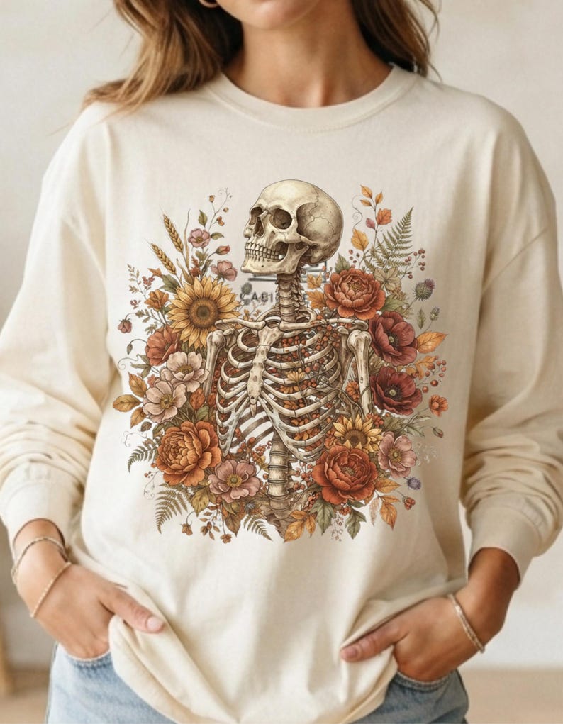 May include: A cream-colored long-sleeve sweatshirt featuring a central graphic of a human skeleton torso surrounded by a vibrant arrangement of autumn-toned flowers, including sunflowers, poppies, and ferns in shades of orange, red, and brown.