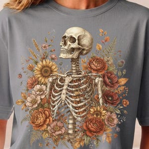 May include: A grey t-shirt featuring a graphic print of a human skeleton torso surrounded by a vibrant arrangement of autumn-toned flowers, including sunflowers, poppies, and peonies, with wheat stalks and fern leaves integrated into the design.