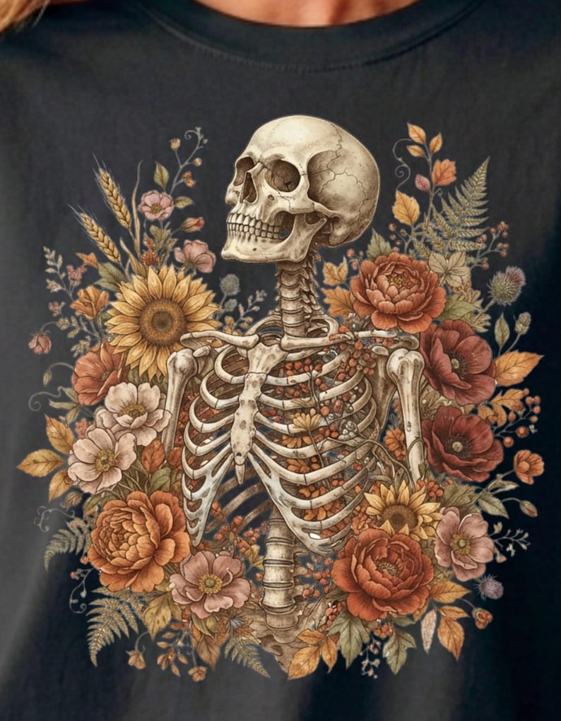 May include: A black t-shirt featuring a detailed graphic of a human skeleton torso and skull surrounded by a vibrant arrangement of autumn-toned flowers, including sunflowers, poppies, and ferns, with small berries nestled within the rib cage.