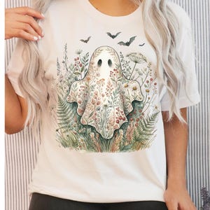 May include: A white t-shirt featuring a central graphic of a ghost covered in a colourful floral pattern. The ghost is surrounded by various wildflowers, ferns, and berries, with several small black bats flying above the ghost in the upper section.