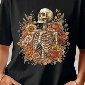 May include: A black t-shirt featuring a graphic print of a human skeleton torso surrounded by a vibrant arrangement of autumn-colored flowers, including sunflowers, poppies, and ferns, with the skeleton facing toward the upper left side.