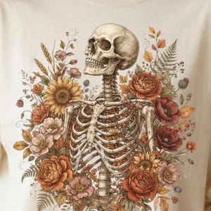 May include: A cream-colored t-shirt featuring a graphic print of a human skeleton torso surrounded by a vibrant arrangement of sunflowers, red poppies, peonies, ferns, and wheat stalks in warm autumn tones of orange, yellow, and deep red.