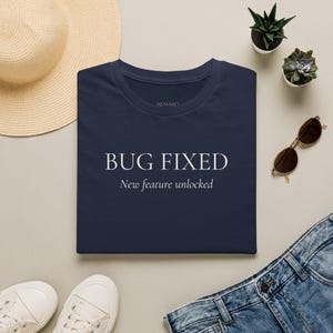 May include: A navy blue t-shirt featuring the white text "BUG FIXED" in a bold serif font, with the smaller white text "New feature unlocked" underneath. The shirt has a crew neckline and the brand name "AEMARO" printed inside the collar area.