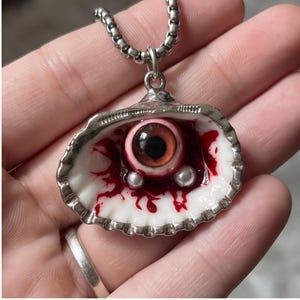 Puede incluir: Un collar con colgante plateado que presenta una concha blanca. Dentro de la concha hay un globo ocular de cristal realista con iris marrón, rodeado de detalles pintados en rojo que simulan sangre y dos pequeñas cuentas esféricas blancas.