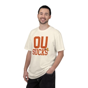 May include: A cream-coloured short-sleeve t-shirt featuring the orange text OU SUCKS printed on the front. A small graphic of a brown cowboy hat is positioned next to the word SUCKS. The shirt has a crew neckline and a standard fit design.