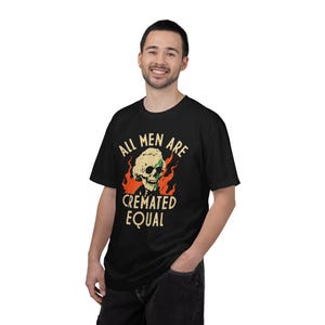 Puede incluir: Una camiseta negra de manga corta con un gráfico de una calavera con peluca rodeada de llamas naranjas. El texto impreso en color amarillo claro dice ALL MEN ARE CREMATED EQUAL, situado encima y debajo de la ilustración central de la calavera.