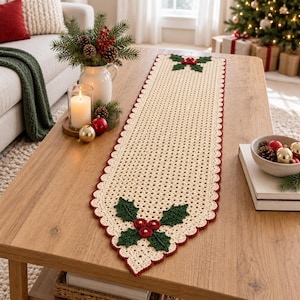 Puede incluir: Un camino de mesa rectangular de ganchillo color crema con un borde festoneado rojo oscuro y motivos decorativos de hojas de acebo con bayas rojas en ambos extremos. El intrincado patrón calado crea una superficie texturizada en el camino.