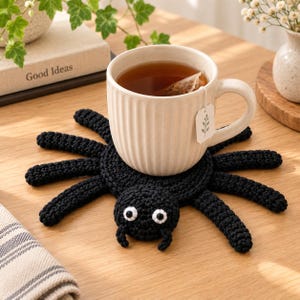 May include: A black crocheted spider coaster with eight legs and two white and black eyes, supporting a ribbed cream-coloured ceramic mug filled with tea and a tea bag tag featuring a small leaf illustration hanging from the side of the mug.