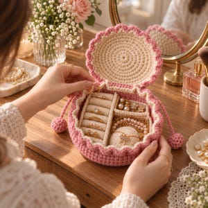 May include: A round, crocheted jewelry box in soft pink and cream yarn with a scalloped edge and drawstring closure. The interior features a ring holder section and a compartment for necklaces and earrings, containing various gold-toned jewelry pieces.