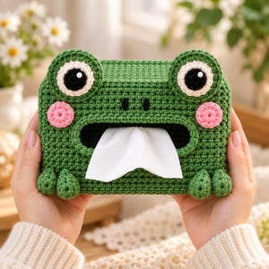 May include: A green crocheted tissue box cover shaped like a frog with large white and black eyes, pink circular cheeks, and a wide mouth opening. A white tissue is pulled through the center of the frog-shaped cover, which features small feet.