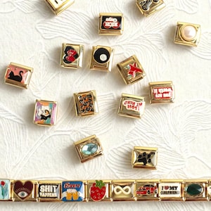 May include: A collection of gold-tone modular bracelet links featuring various enamel designs, including a black cat, an eight ball, a strawberry, a pearl, and text such as "SHIT HAPPENS" and "I LOVE MY GIRLFRIEND," arranged with a connected bracelet.