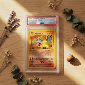 May include: A 1999 French Charizard holographic trading card encased in a clear protective slab. The card features a yellow border, a central illustration of the orange dragon-like creature, and text including 120 HP and Fire Spin attack details.